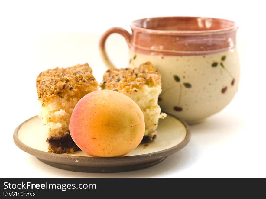 A cup of milk and a plate on which lay pieces of cake and a ripe apricot, on a white background. A cup of milk and a plate on which lay pieces of cake and a ripe apricot, on a white background