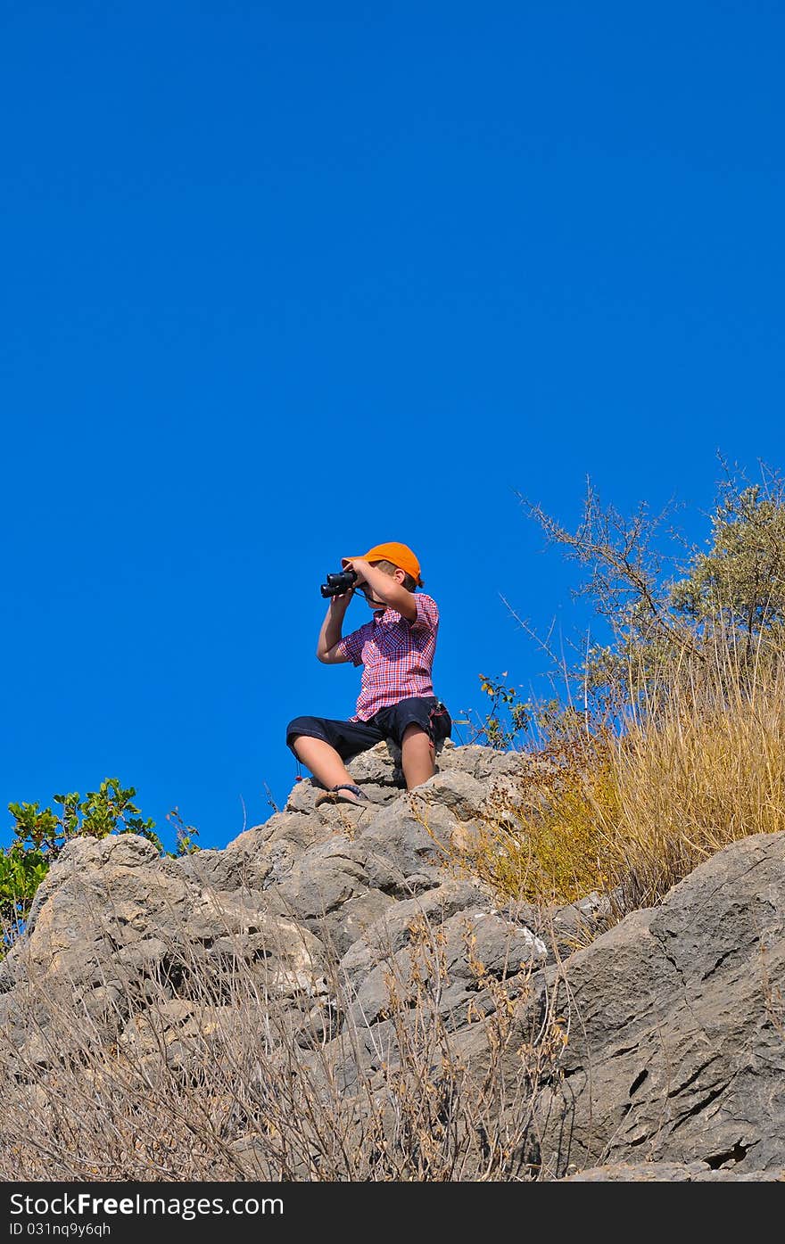 Boy sitting on a rock on top of a mountain and sees the neighborhood through binoculars. Boy sitting on a rock on top of a mountain and sees the neighborhood through binoculars