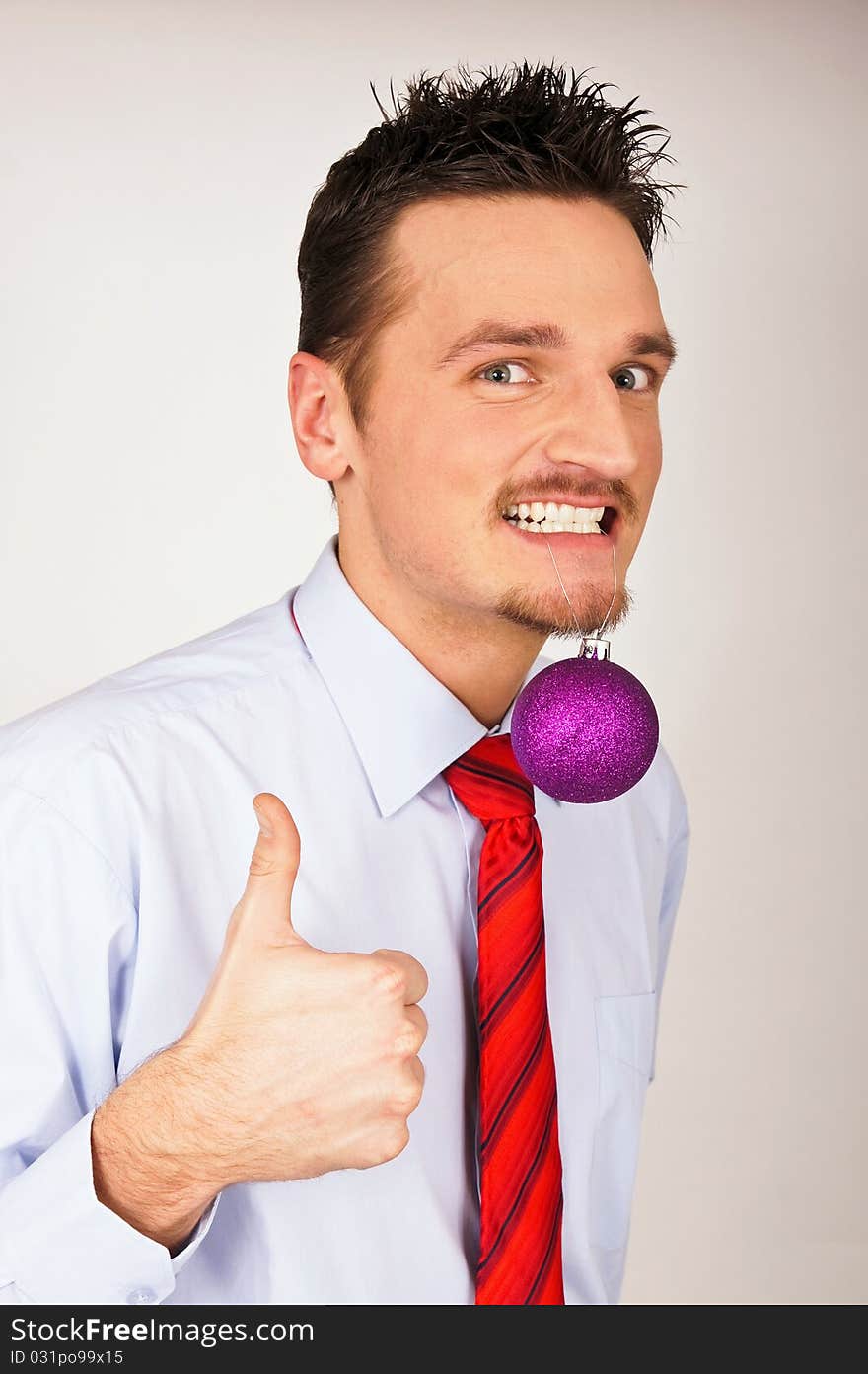 Happy young man in shirt and tie has violet Christmas Ornament between teeth and shows with thumb yes. Happy young man in shirt and tie has violet Christmas Ornament between teeth and shows with thumb yes.