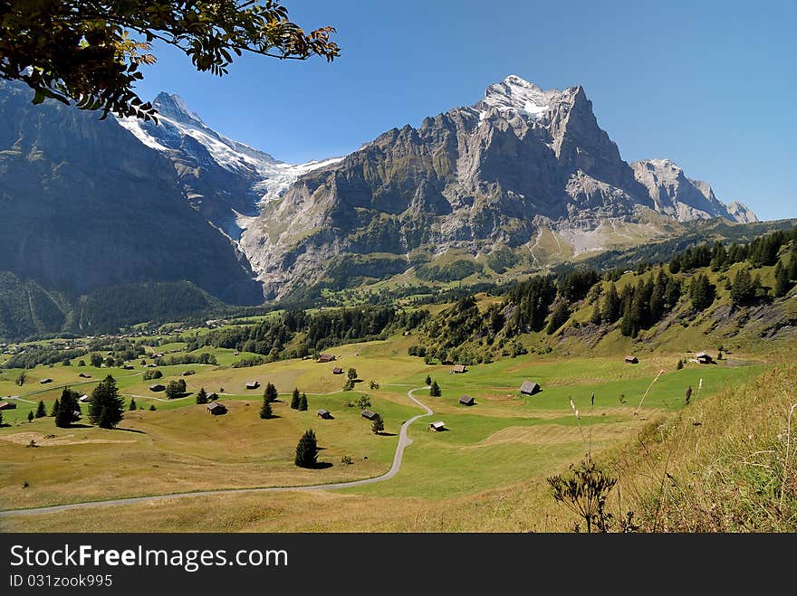 The road across an alpine village towards the high mountains with glaciers highlighted with sun, against the background of the blue sky. The road across an alpine village towards the high mountains with glaciers highlighted with sun, against the background of the blue sky