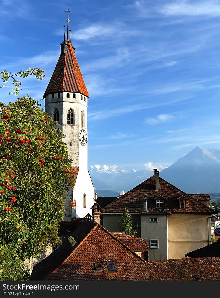 The clock tower with red tiled roof raising over the buildings of the old city, against the background of bright blue sky, with a view of distant mountains