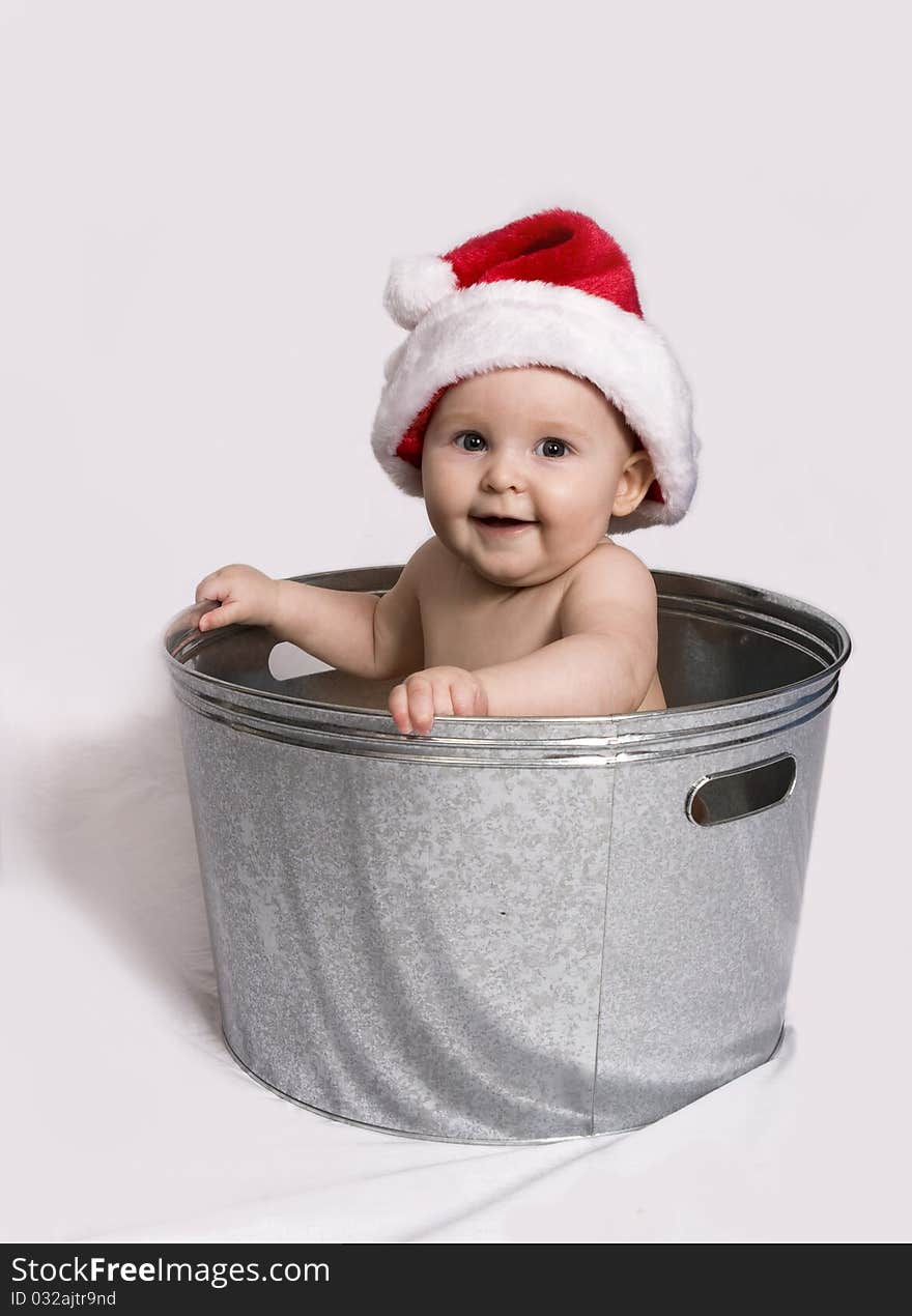 Smiling baby wearing a Santa hat, holds on to the sides of a wash basin while sitting in it. Smiling baby wearing a Santa hat, holds on to the sides of a wash basin while sitting in it.