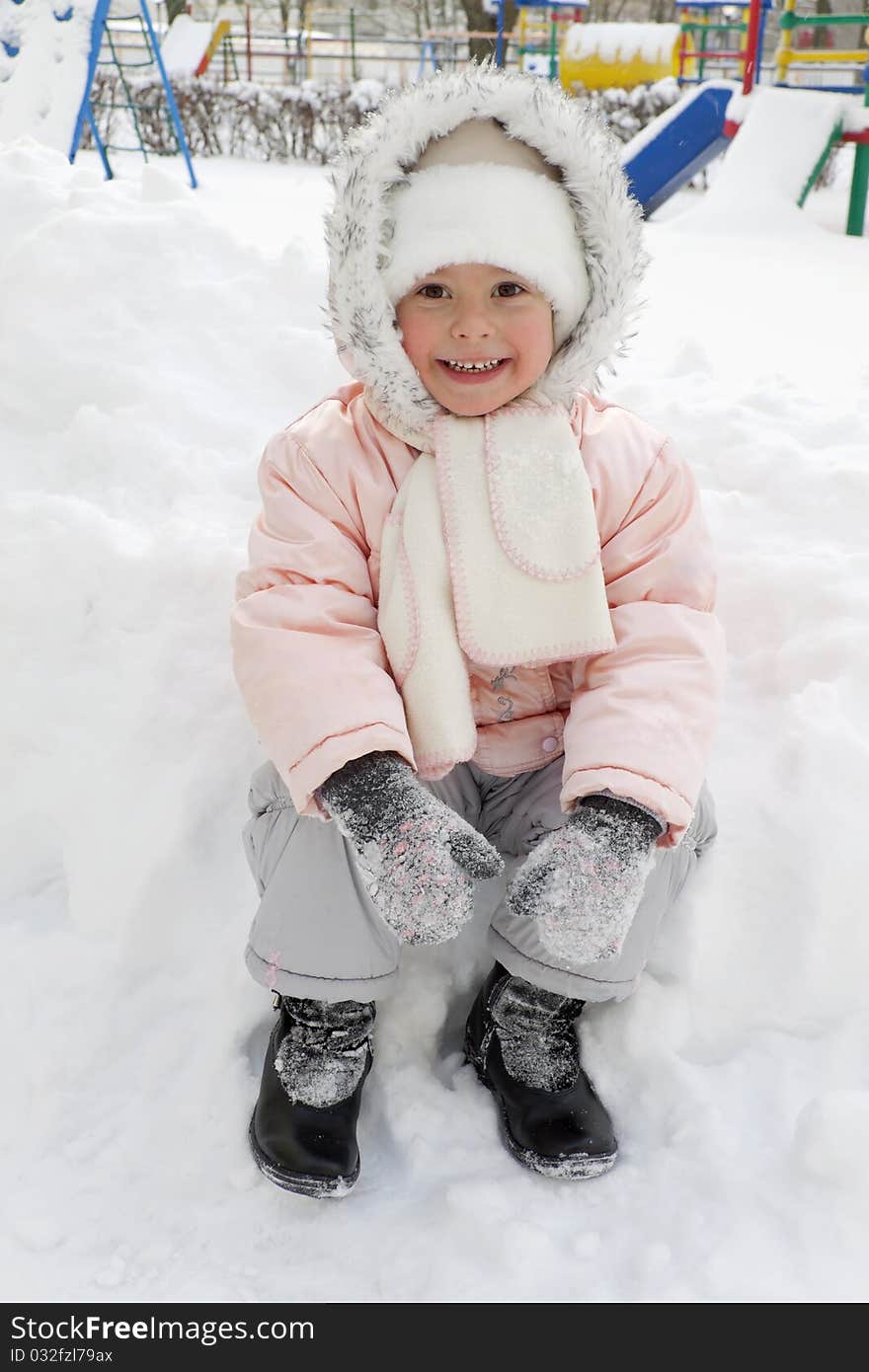 Warmly dressed joyful child sits in a snowdrift on a playground. Warmly dressed joyful child sits in a snowdrift on a playground