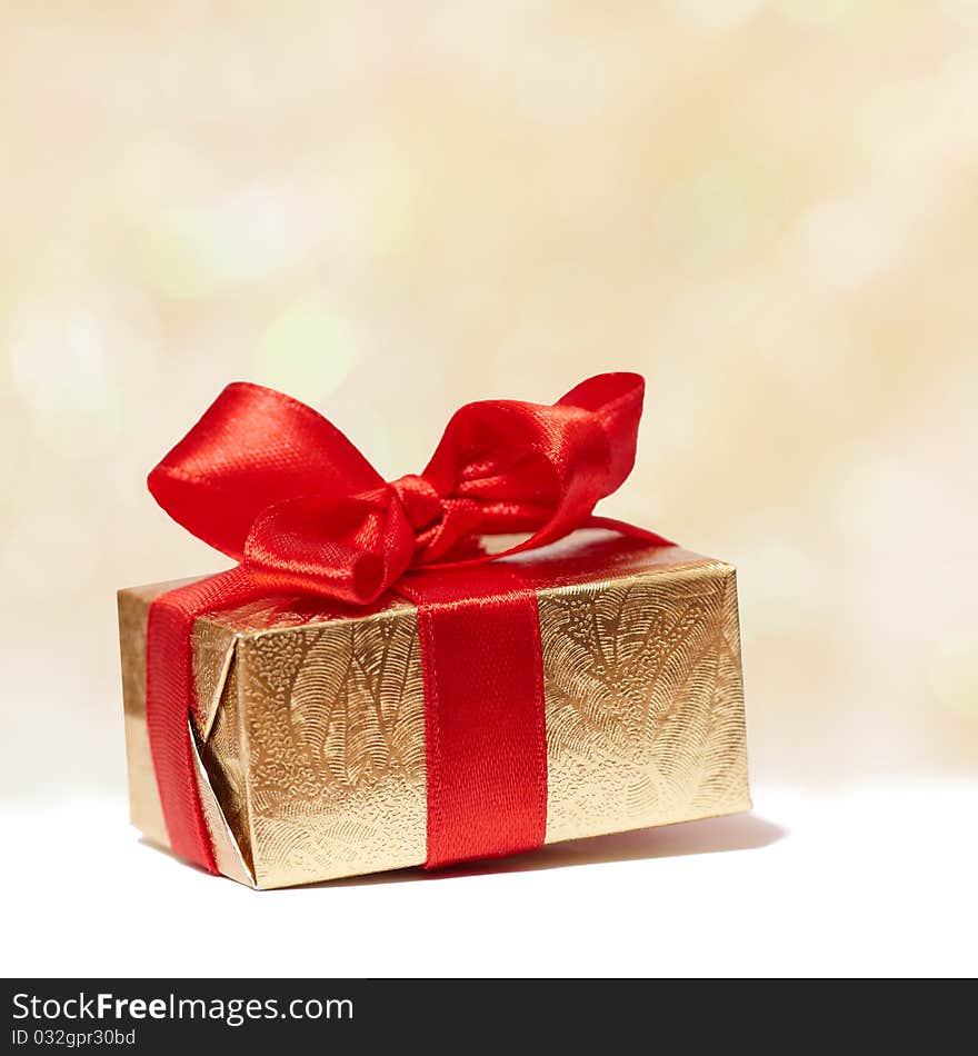 Golden christmas gift box with red ribbon and bow on background of defocused golden lights. Golden christmas gift box with red ribbon and bow on background of defocused golden lights