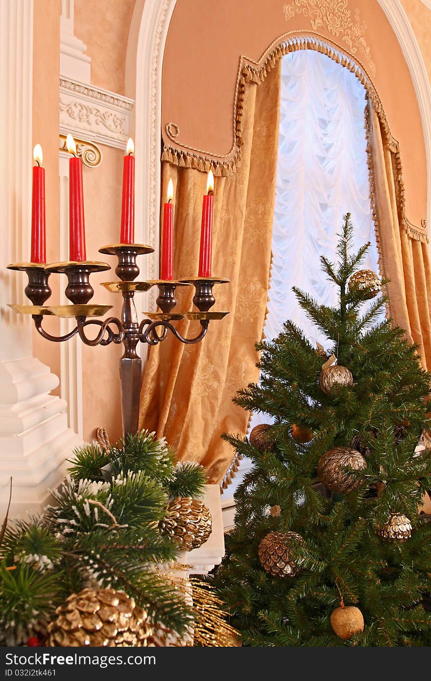 Candles in a candlestick on a fireplace against a window with beautiful portieres and a Christmas fur-tree.