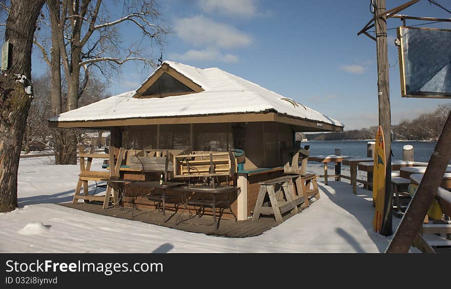 Small beach shack sits next the water, boarded up and closed for the season. It is snow covered with tables and chairs stacked on its deck against a bright blue winter sky. Small beach shack sits next the water, boarded up and closed for the season. It is snow covered with tables and chairs stacked on its deck against a bright blue winter sky.