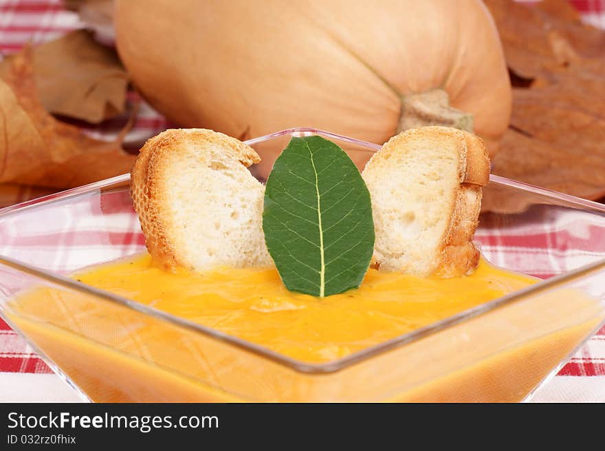 Cream of squash soup in a squared glass plate with toasted bread and a bay leaf. Pumpkin out of focus in the background. Studio shot. Selective focus, shallow DOF.