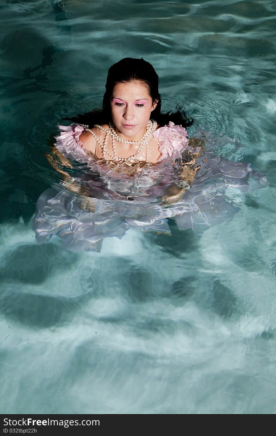 A woman standing in water in her fancy pink dress with her pearl necklace on with a peaceful expression on her face. A woman standing in water in her fancy pink dress with her pearl necklace on with a peaceful expression on her face.