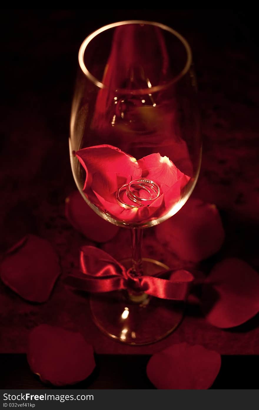 A pair of silver wedding ring in a red wine glass with fresh red rose petals underneath it. A pair of silver wedding ring in a red wine glass with fresh red rose petals underneath it.