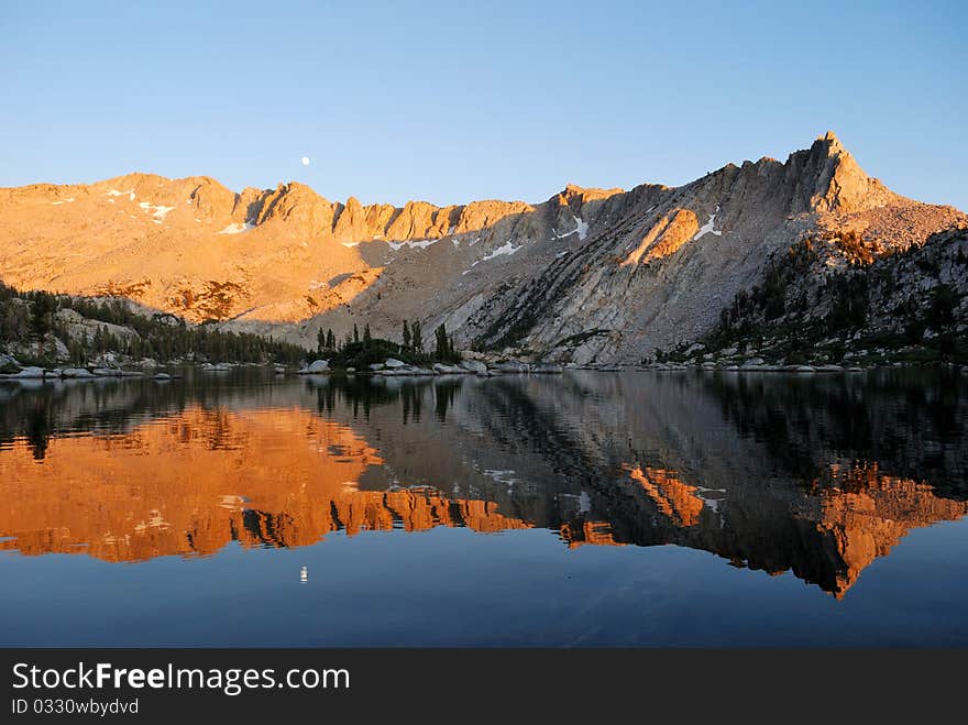 Moon rising over a mountain ridge glowing orange from the setting sun, reflected in a calm alpine lake. Moon rising over a mountain ridge glowing orange from the setting sun, reflected in a calm alpine lake.