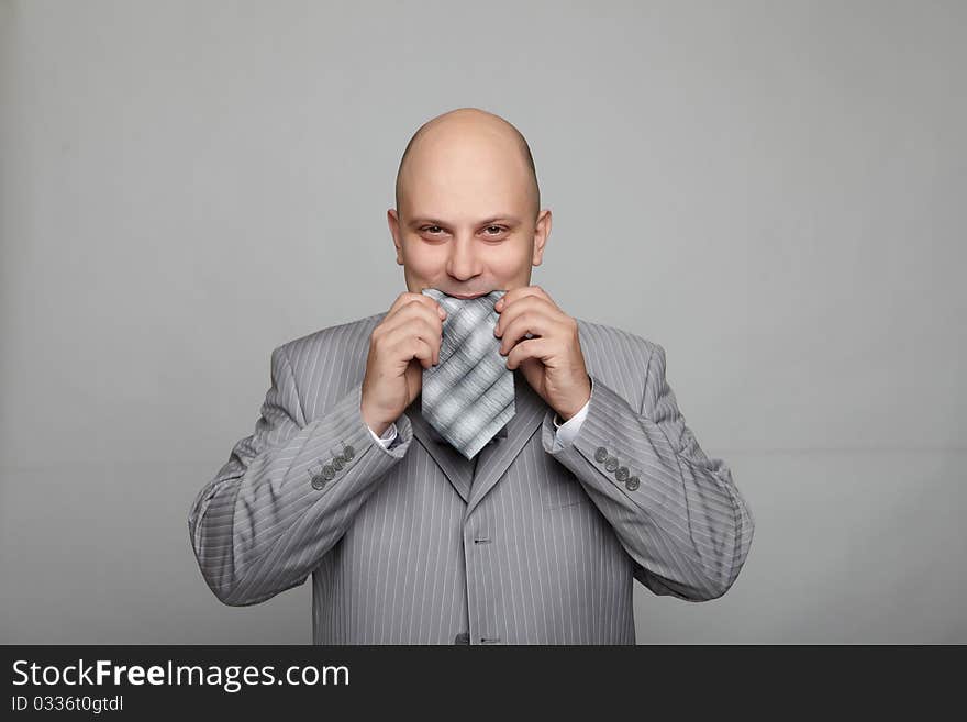 Bald businessman in a gray suit with a gray background eats his own tie. Funny business