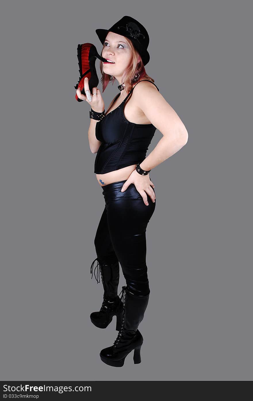 A young punk woman in black leather pants, black corset and boots and a hat standing with her back, for gray background. A young punk woman in black leather pants, black corset and boots and a hat standing with her back, for gray background.