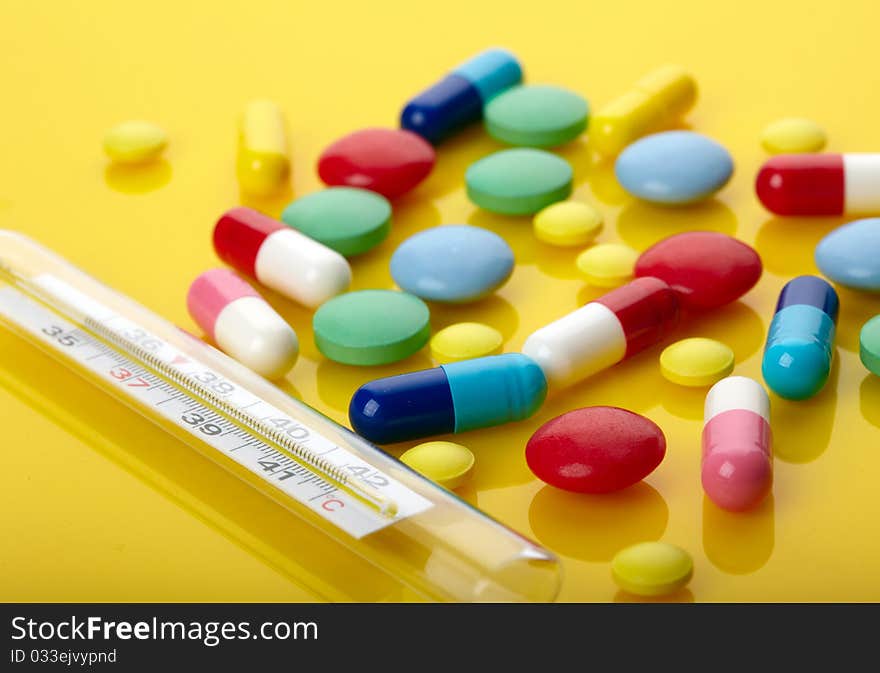 Colorful pills and thermometer over yellow background