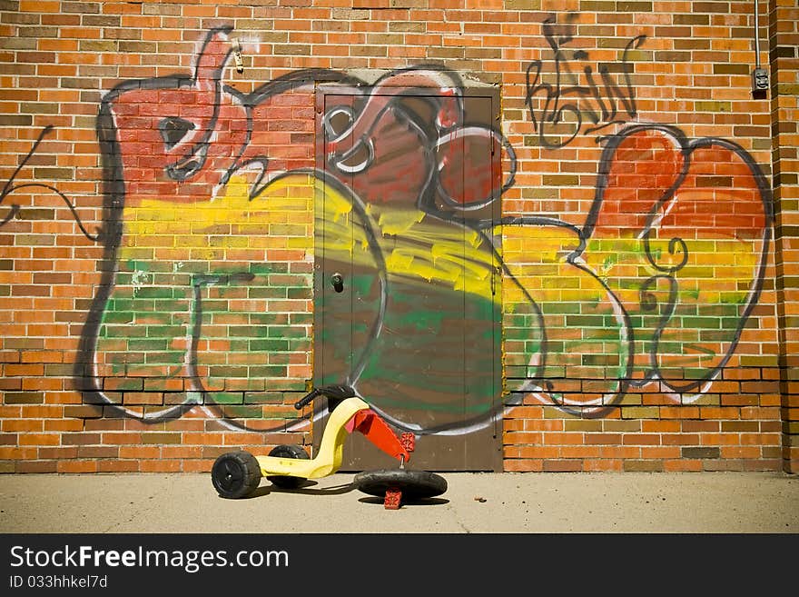 A broken child's tricycle is abandoned near an old storage warehouse that has been vandalized with graffiti. A broken child's tricycle is abandoned near an old storage warehouse that has been vandalized with graffiti.