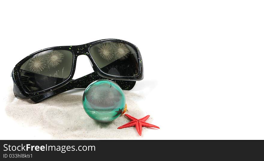 An angled closeup view of a pair of sunglasses, christmas bulb and a red star fish on some white beach sand. Reflections of the caribbean ocean with a parasailer and fireworks in the christmas buld and sunglasses. An angled closeup view of a pair of sunglasses, christmas bulb and a red star fish on some white beach sand. Reflections of the caribbean ocean with a parasailer and fireworks in the christmas buld and sunglasses.