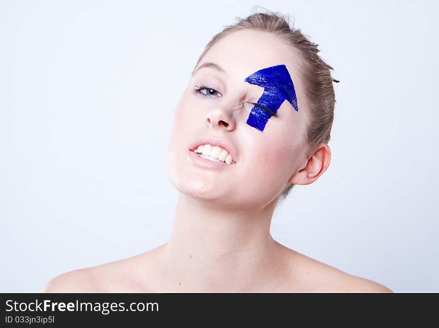 Portrait of smiling young blond haired woman with upward pointing arrow painted over eye, white studio background.