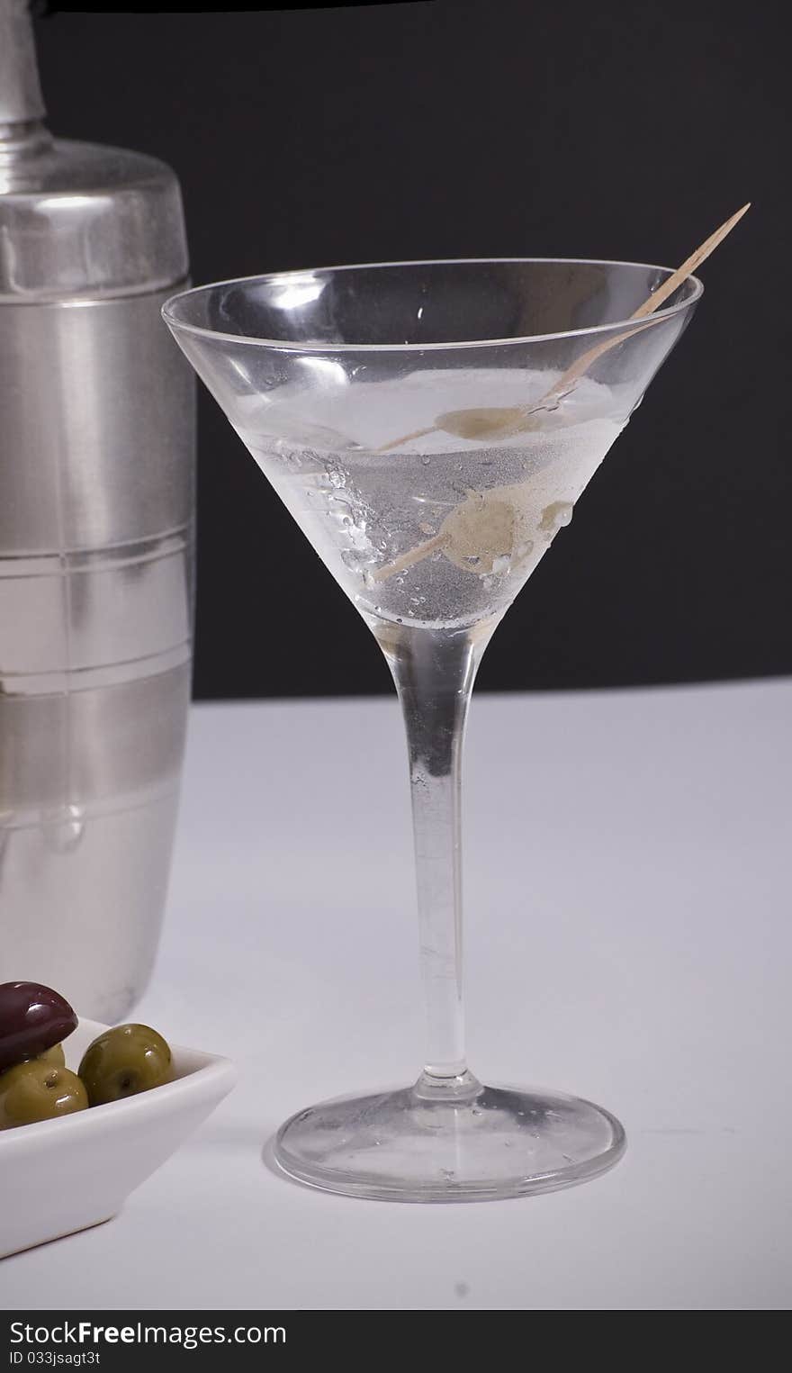 A glass of ice cold martini with cocktail shaker and olives in background