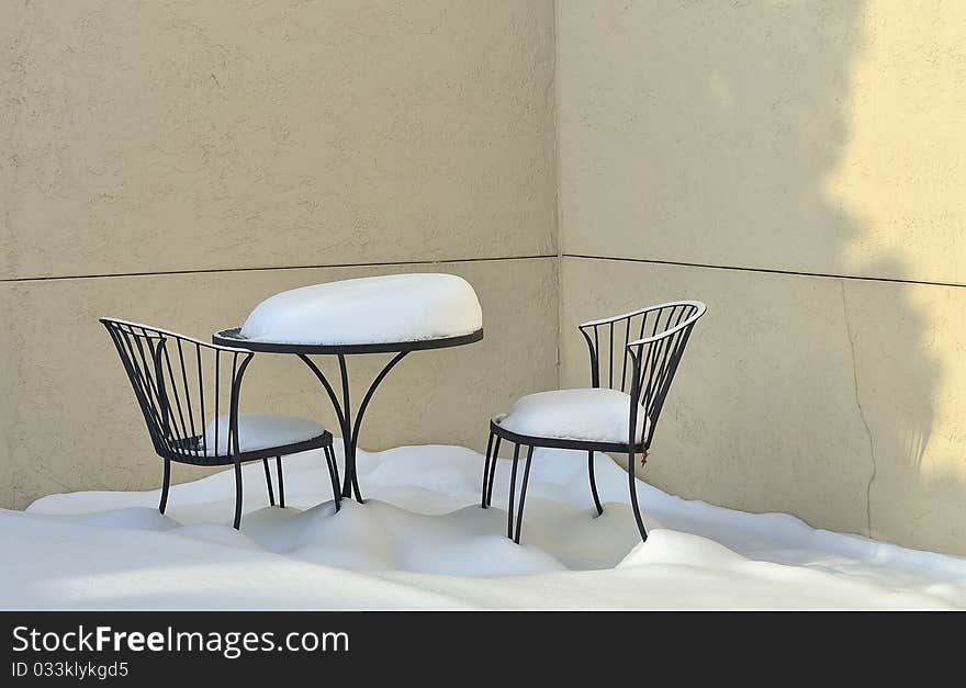 Patio table and two chairs outdoors on a empty patio with mounds of new fallen snow covering them and the floor. Patio table and two chairs outdoors on a empty patio with mounds of new fallen snow covering them and the floor.