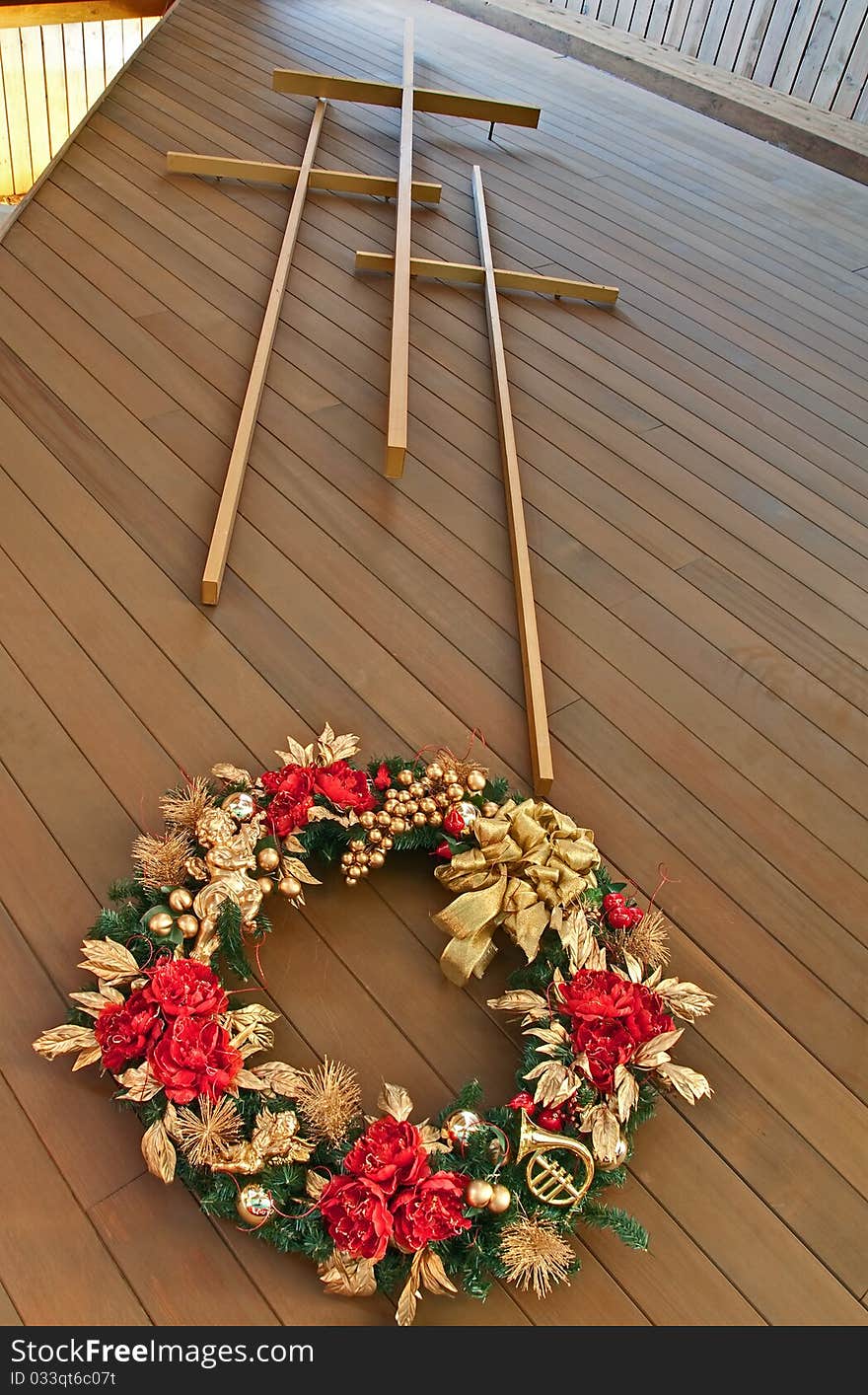 This stock image has three crosses with a view of looking up at the crosses from the foot of the cross inside a portion of a church. There is a gold and red Christmas wreath hanging beneath the crosses. This stock image has three crosses with a view of looking up at the crosses from the foot of the cross inside a portion of a church. There is a gold and red Christmas wreath hanging beneath the crosses.