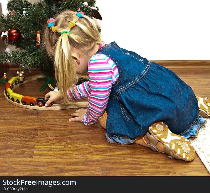 Blonde girl in jeans sarafan playing with toy wooden train, toys under the fir tree, copy space. Blonde girl in jeans sarafan playing with toy wooden train, toys under the fir tree, copy space