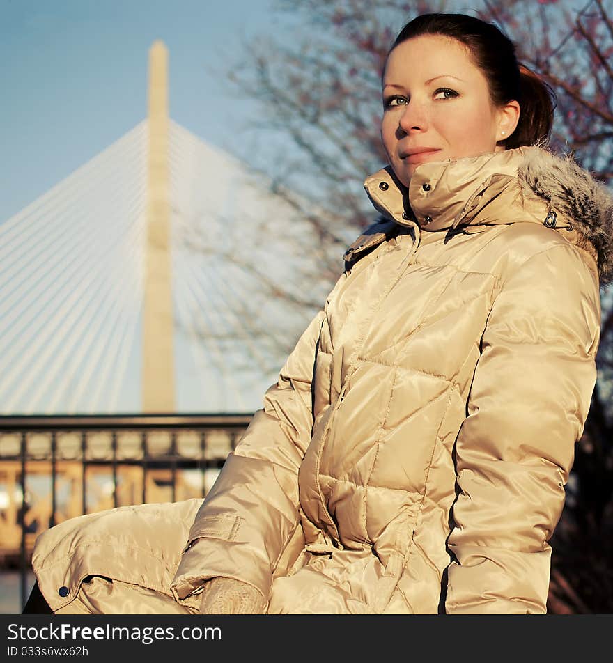 Girl in beige dawn parka sitting on a bench with a bridge on the background, Boston