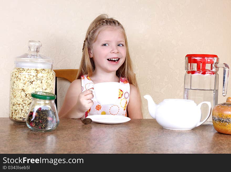 Little girlwith cup of hot drink, at table with tea, jasmin petals and teapot against yellow wall in home. Little girlwith cup of hot drink, at table with tea, jasmin petals and teapot against yellow wall in home