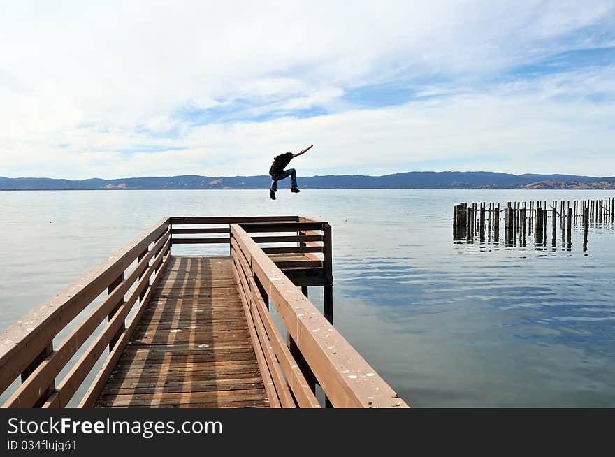 A man jumping a long narrow wooden pier into the calm blue ocean water with outstretched victorious arms. A man jumping a long narrow wooden pier into the calm blue ocean water with outstretched victorious arms.