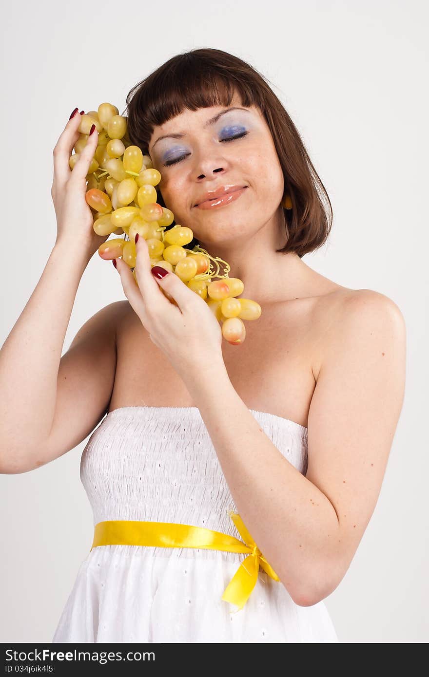 Beautiful girl in white dress with yellow tape is holding green grapes. She is isolated on a white background. Beautiful girl in white dress with yellow tape is holding green grapes. She is isolated on a white background.