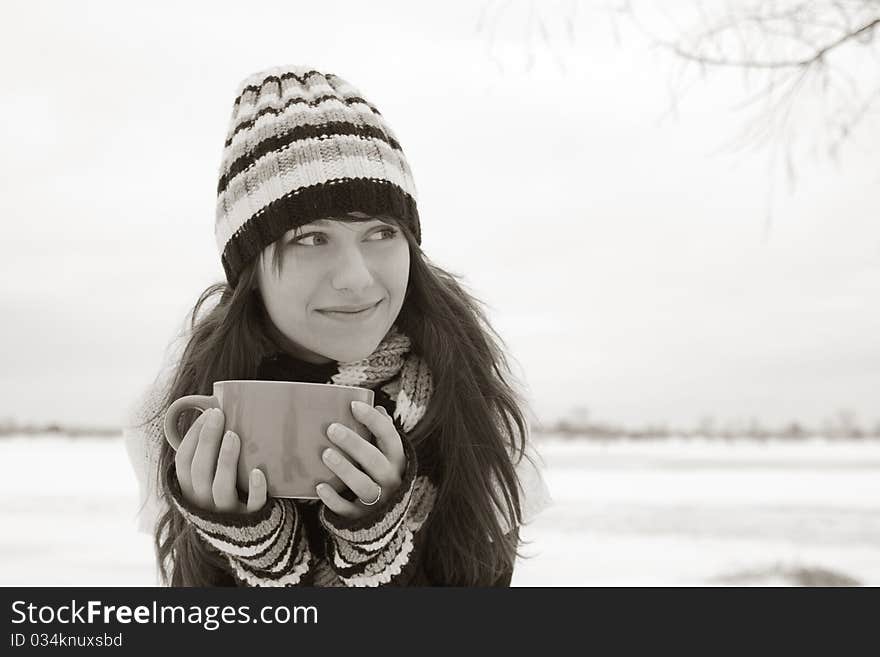 Young beautiful smiling woman in a striped hat and mittens sitting on the street with a large cup in his hand. Young beautiful smiling woman in a striped hat and mittens sitting on the street with a large cup in his hand