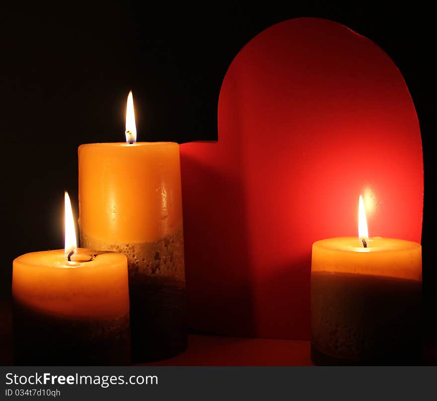 Lit Candles are illuminating a red love heart on a black background. Lit Candles are illuminating a red love heart on a black background