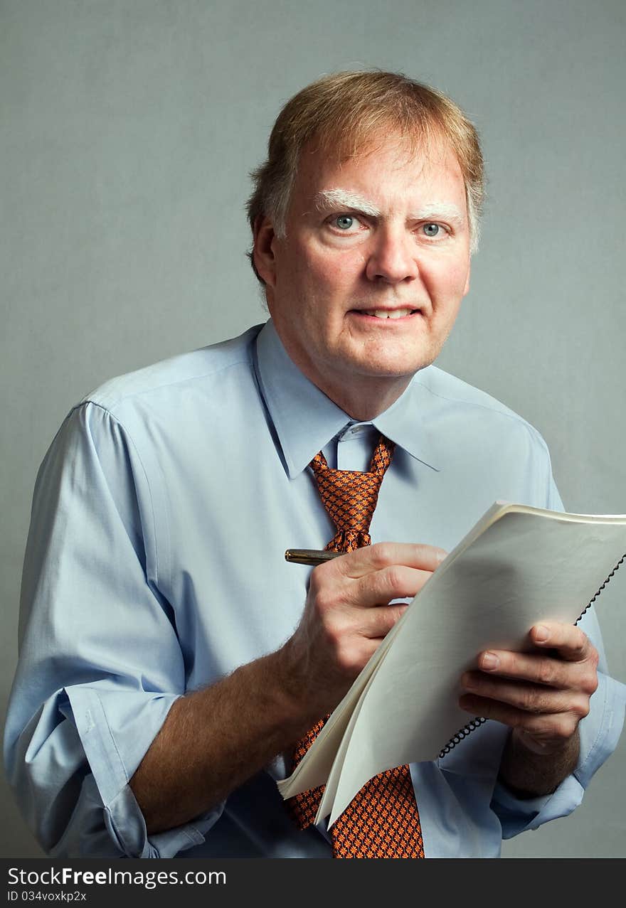 Businessman, professional, writing on paper, smiling and looking at the camera. The man is wearing a blue shirt and orange necktie and is in front of a white background. His shirt sleeves are rolled up and collar loosened. Businessman, professional, writing on paper, smiling and looking at the camera. The man is wearing a blue shirt and orange necktie and is in front of a white background. His shirt sleeves are rolled up and collar loosened.