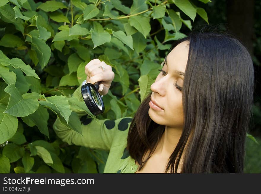 Close-up of an attractive young woman outdoors looking through a magnifying glass on the green leaves of the tree. Close-up of an attractive young woman outdoors looking through a magnifying glass on the green leaves of the tree