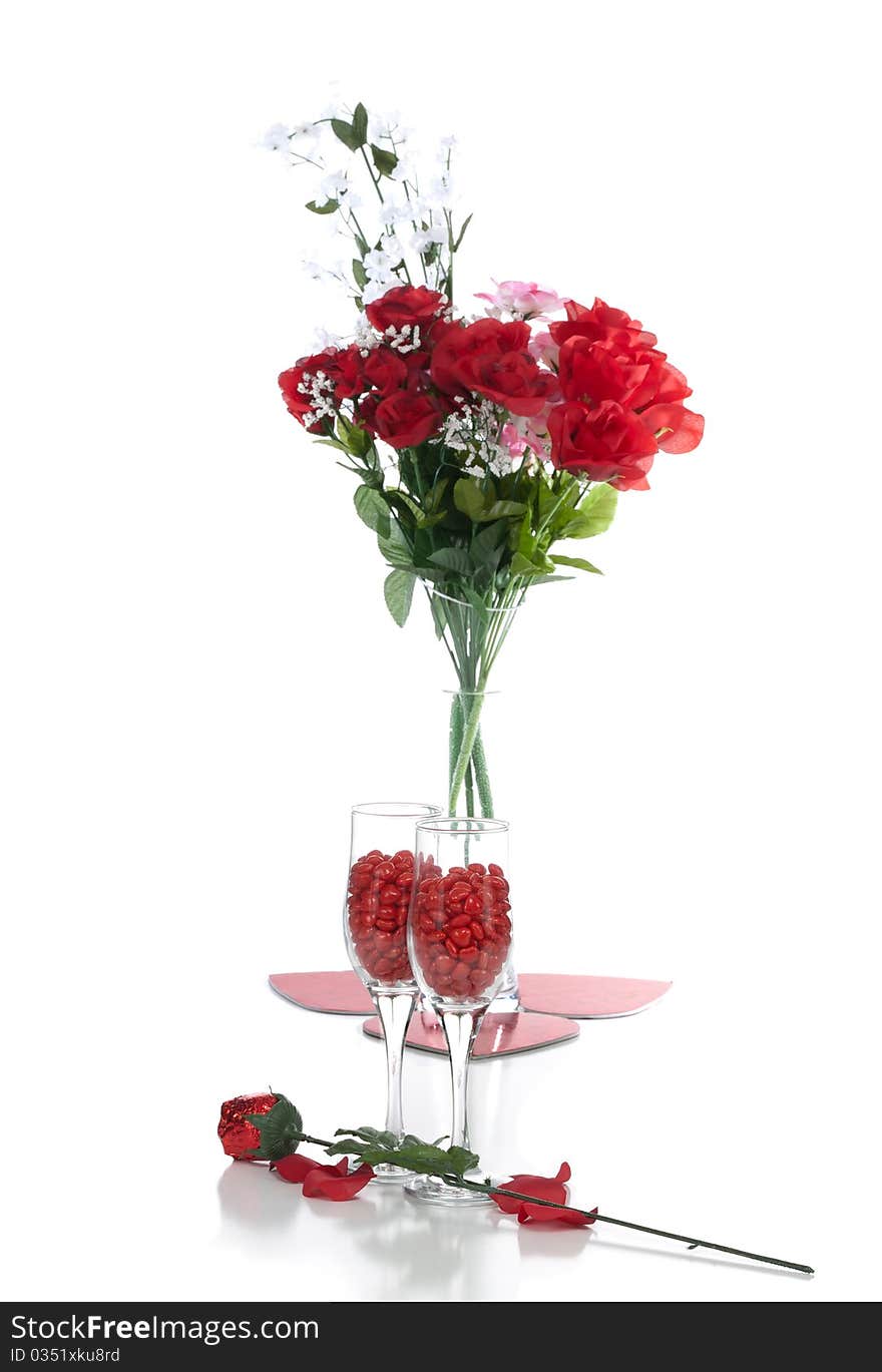 A front isolated studio view of two fluted wine glasses containing cinnamon hearts candy and a red chocolate rose in the foreground ; a vase of fabric flowers and heart shape costers in the background. A front isolated studio view of two fluted wine glasses containing cinnamon hearts candy and a red chocolate rose in the foreground ; a vase of fabric flowers and heart shape costers in the background.