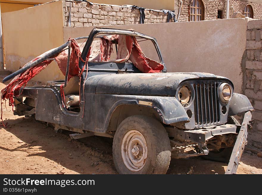 Old jeep without a bumper and without the rear wheels under a layer of dust and sand covered with the remains of the red carpet. Old jeep without a bumper and without the rear wheels under a layer of dust and sand covered with the remains of the red carpet.