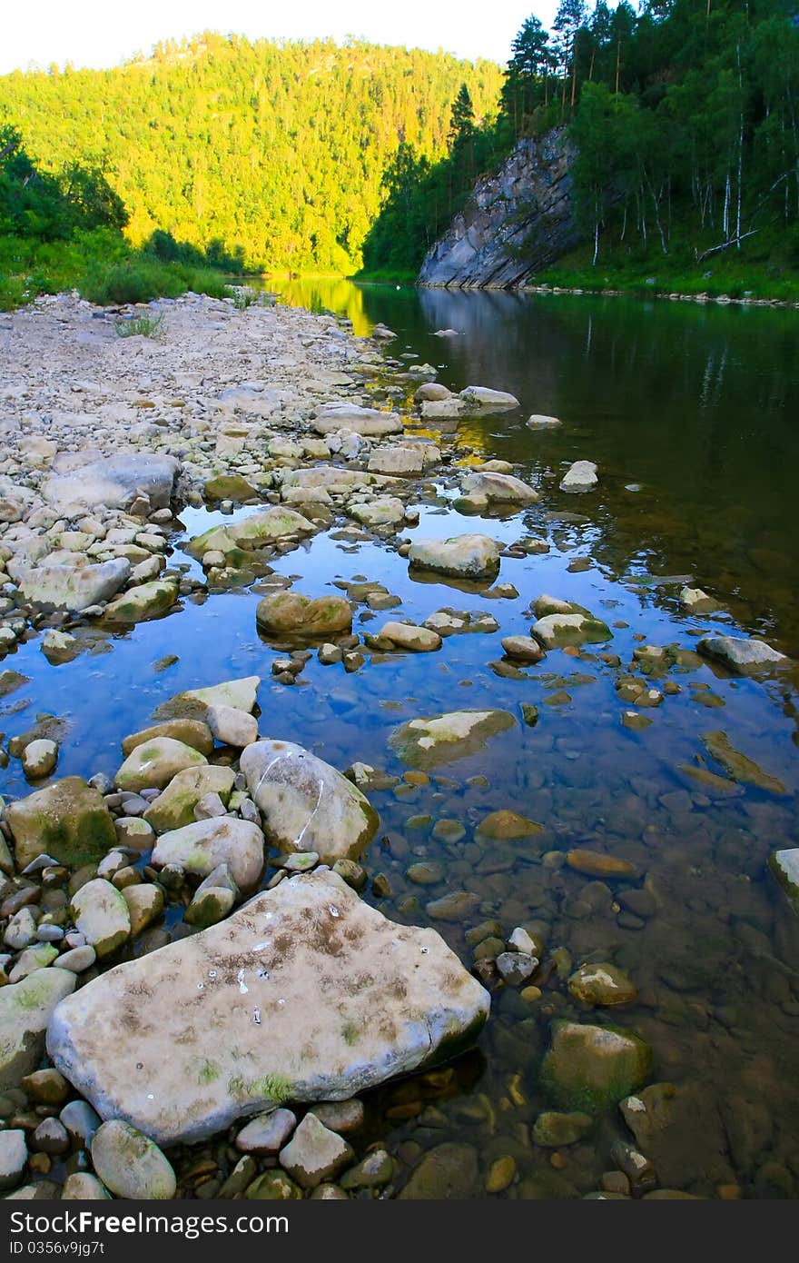 River in the evening. Bright sunlight reflected on a distant hill, covered with forests. Stones in the water in the foreground