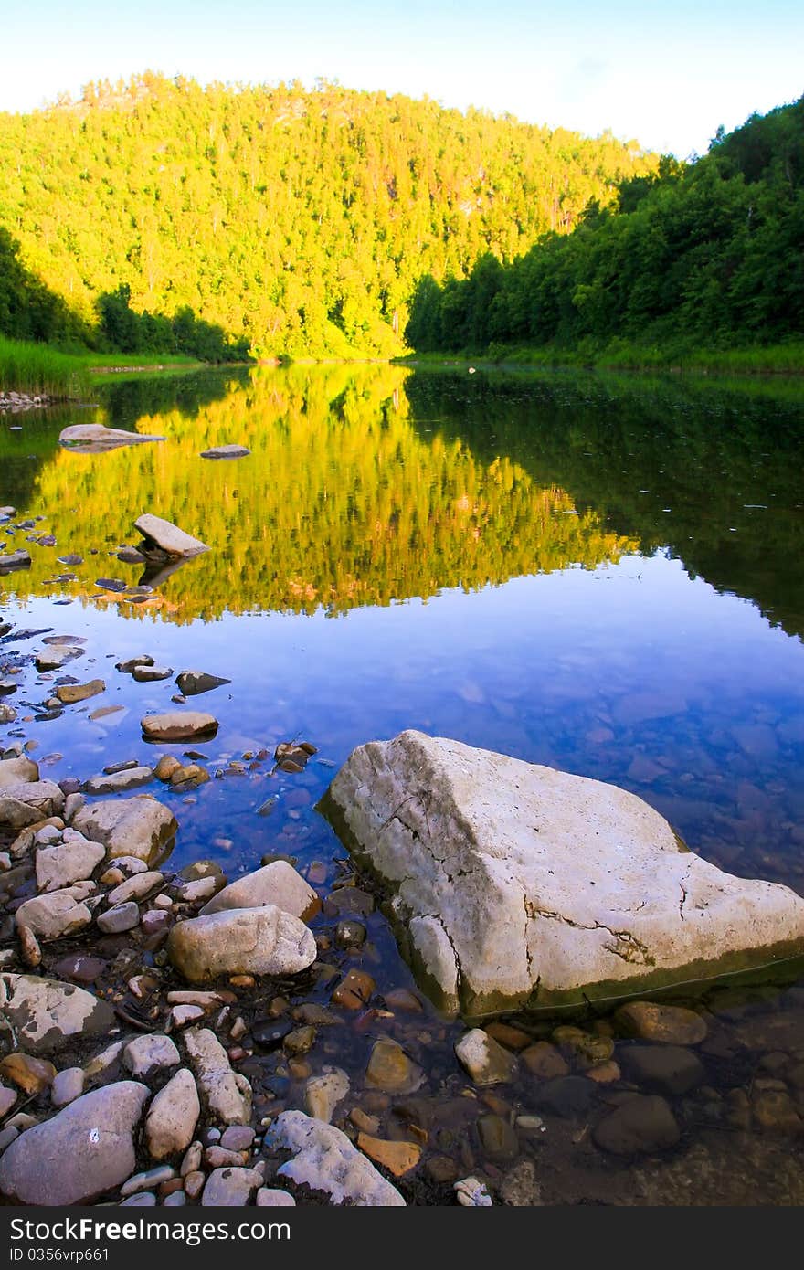 River in the evening. Bright sunlight reflected on a distant hill, covered with forests. Stones in the water in the foreground