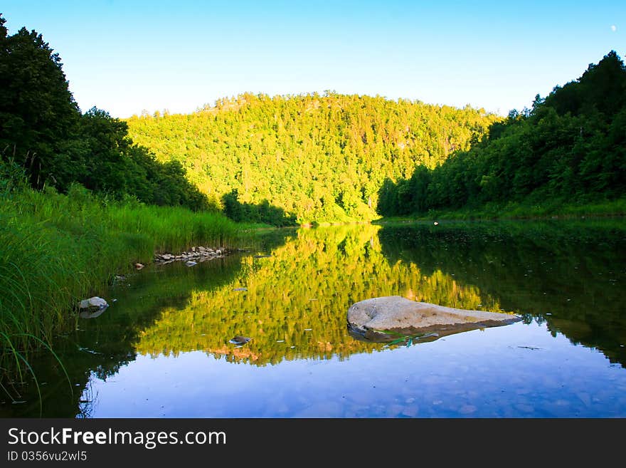 River in the evening. Bright sunlight reflected on a distant hill, covered with forests. Stones in the water in the foreground