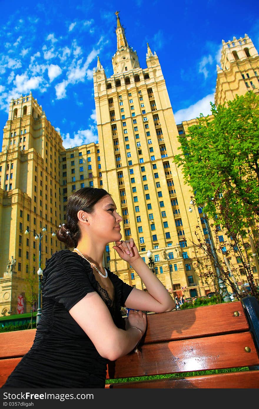 Beautiful Asian girl in black dress sitting on a bench on a lovely day on the background of high beautiful building. Beautiful Asian girl in black dress sitting on a bench on a lovely day on the background of high beautiful building