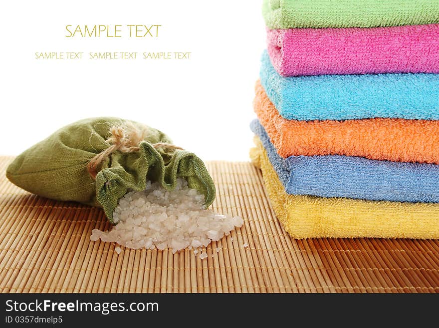 On a wooden mat lots of colorful bath towels stacked on each other lies near the fabric bag filled with bath salts. On a wooden mat lots of colorful bath towels stacked on each other lies near the fabric bag filled with bath salts.