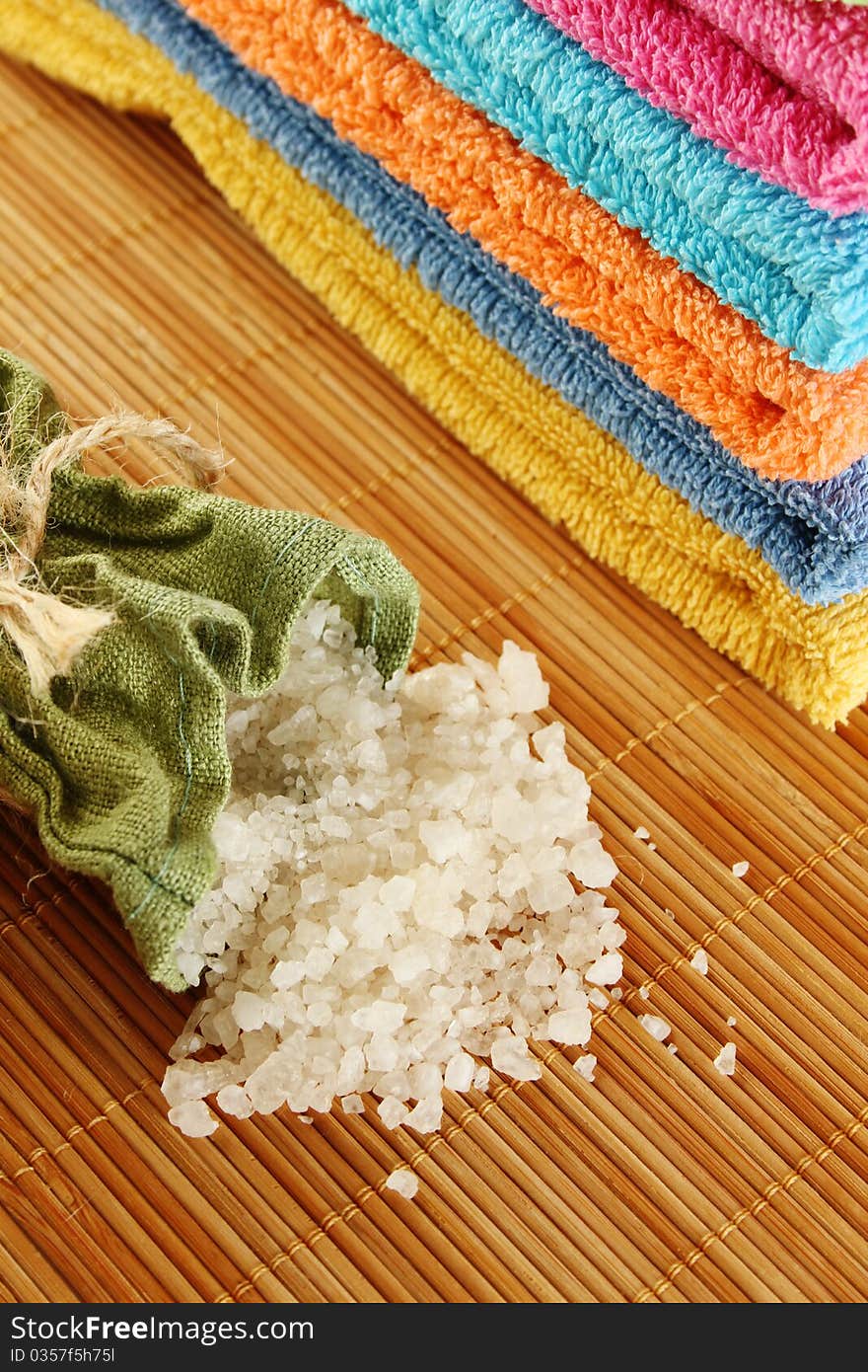 On a wooden mat lots of colorful bath towels stacked on each other lies near the fabric bag filled with bath salts. On a wooden mat lots of colorful bath towels stacked on each other lies near the fabric bag filled with bath salts.