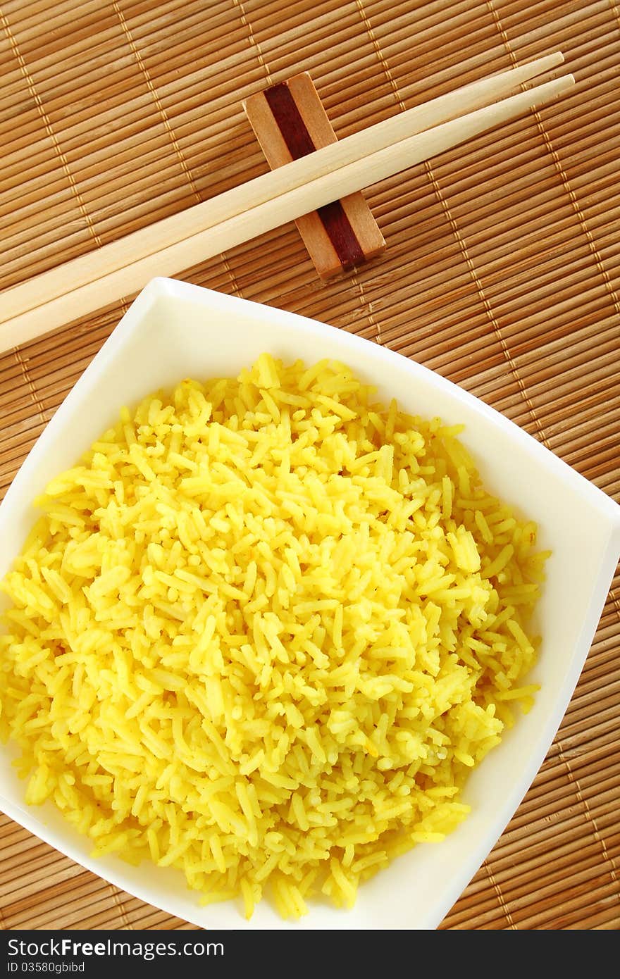 Boiled yellow rice and a white plate beside lie wooden sticks. Plate stands on a wooden mat. Boiled yellow rice and a white plate beside lie wooden sticks. Plate stands on a wooden mat