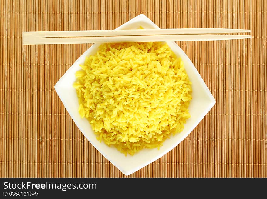 Boiled yellow rice and a white plate beside lie wooden sticks. Plate stands on a wooden mat. Boiled yellow rice and a white plate beside lie wooden sticks. Plate stands on a wooden mat