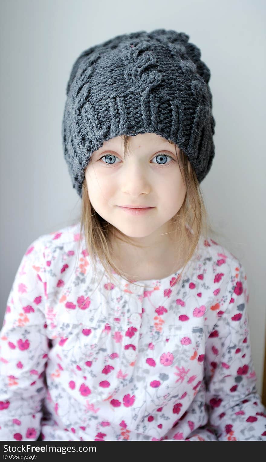 Adorable little kid girl with blue eyes in dark grey knit hat near the window looks into the camera. Adorable little kid girl with blue eyes in dark grey knit hat near the window looks into the camera