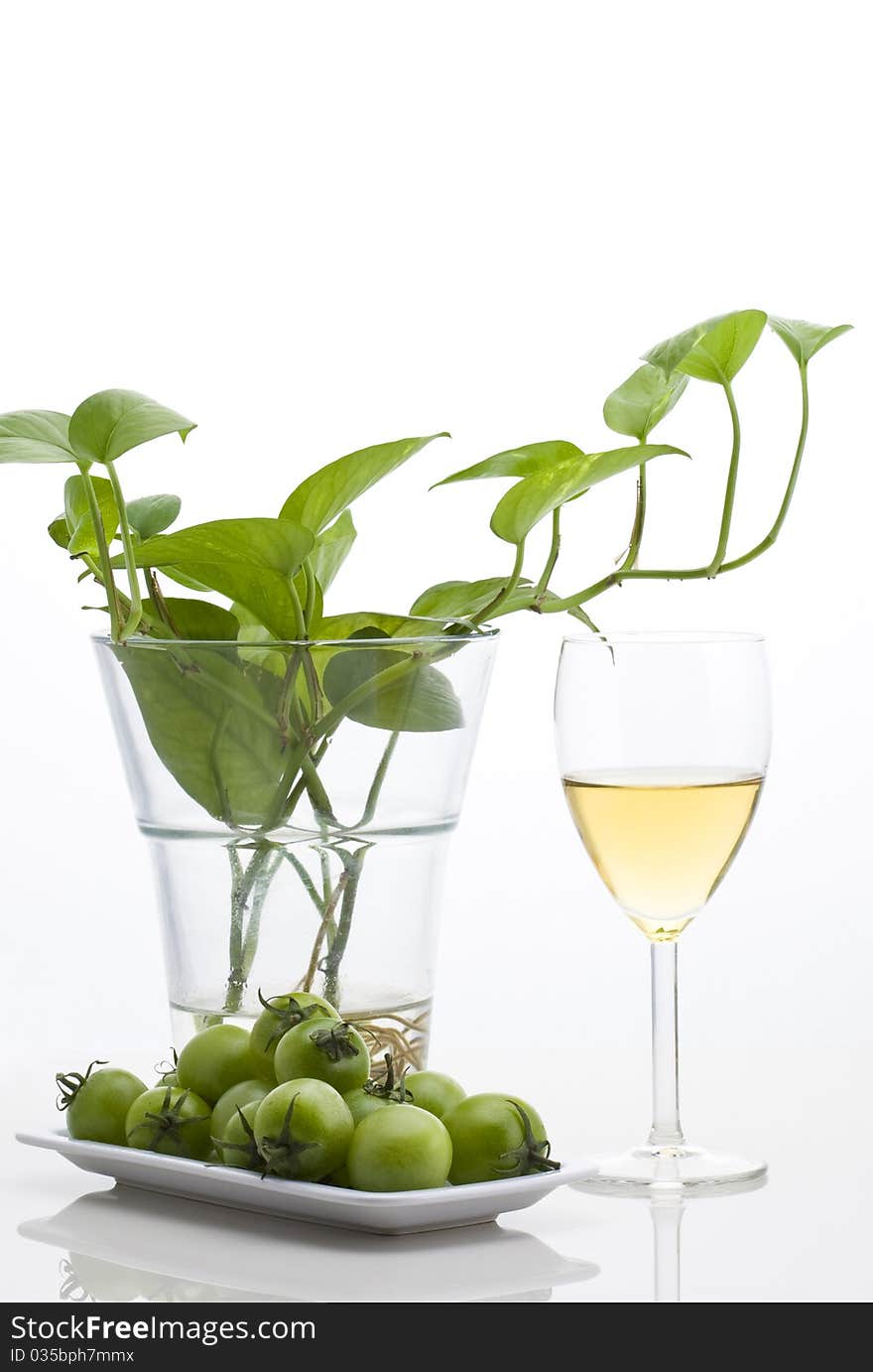 Glass of white wine with green plants and small green tomato on white background.