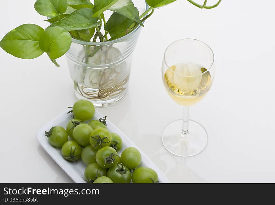 Glass of white wine with green plants and small green tomato on white background.