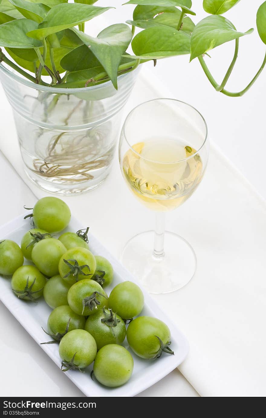 Glass of white wine with green plants and small green tomato on white background.