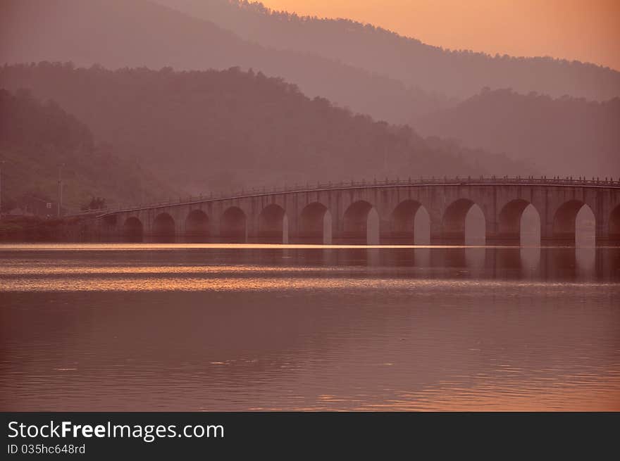 A Chinese ancient stone bridge in the rosy sunset at the mountain foot over the flowing rivulet. A Chinese ancient stone bridge in the rosy sunset at the mountain foot over the flowing rivulet.