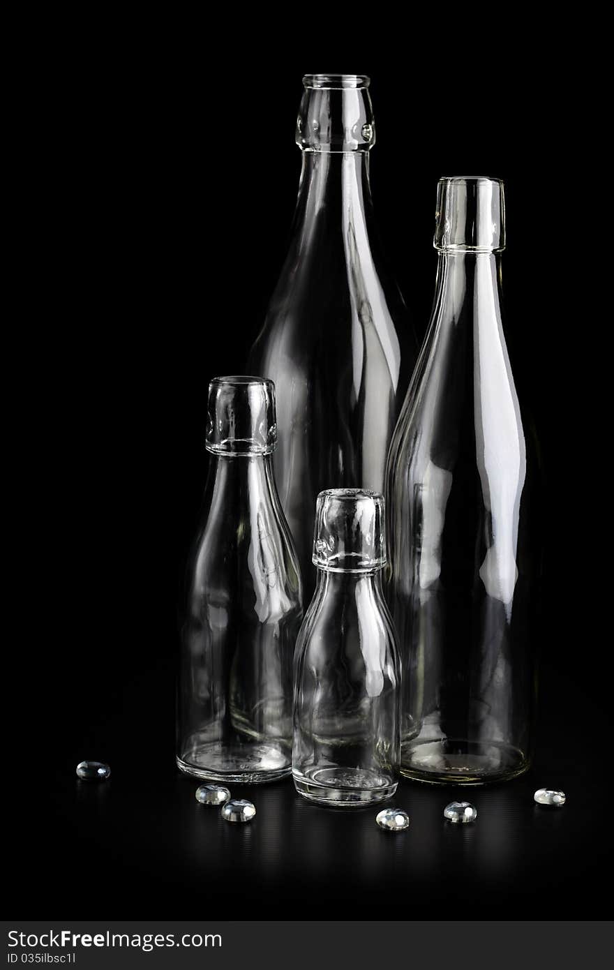 Four empty clear glass bottles and some clear glass pebbles on a black background.