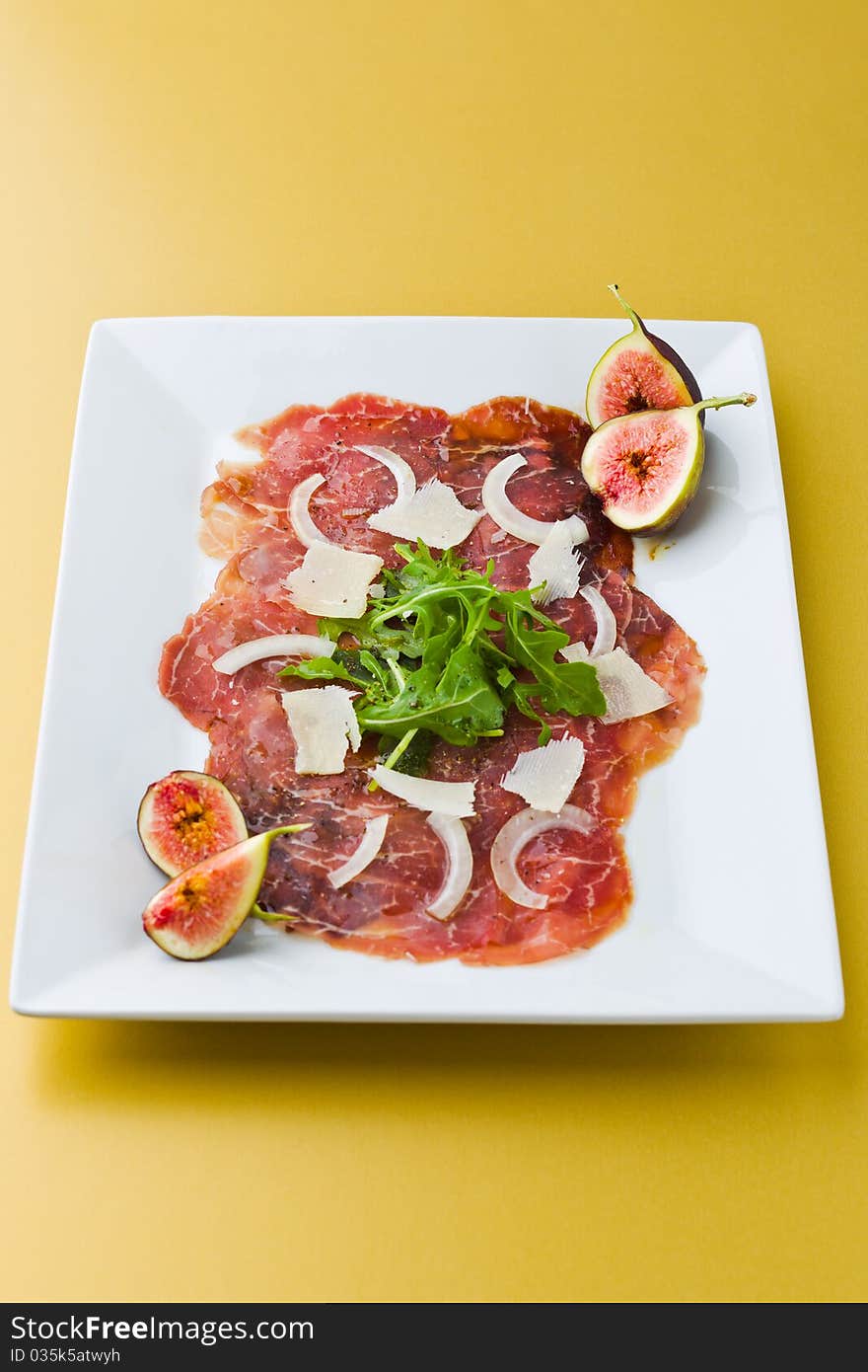 Red beef carpaccio in a white plate with salad, fig fruit, parmesan cheese and olive oil. Yellow background and shallow depth of field. Red beef carpaccio in a white plate with salad, fig fruit, parmesan cheese and olive oil. Yellow background and shallow depth of field.