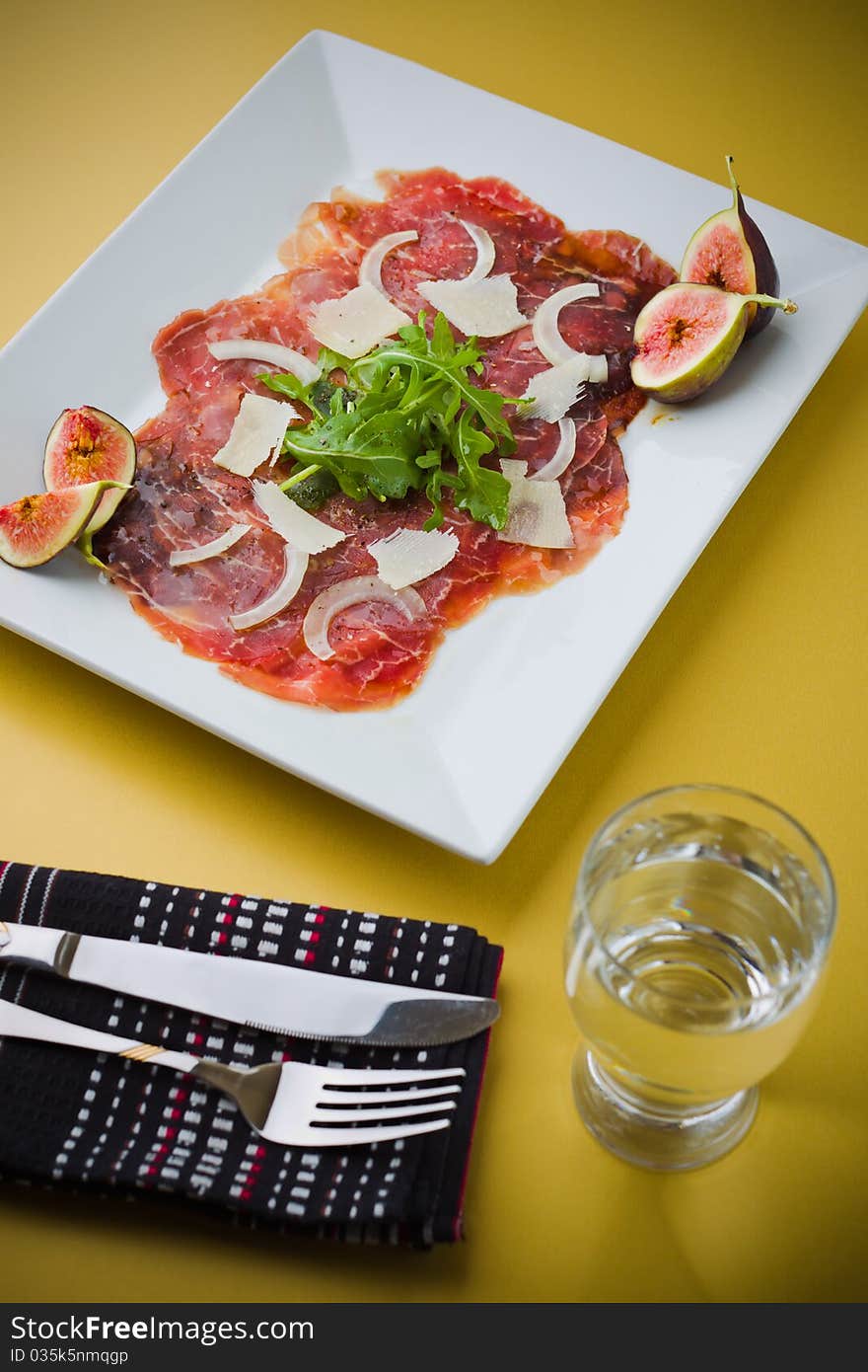 Red beef carpaccio in a white plate with salad, fig fruit, parmesan cheese and olive oil. Yellow background and shallow depth of field. Red beef carpaccio in a white plate with salad, fig fruit, parmesan cheese and olive oil. Yellow background and shallow depth of field.